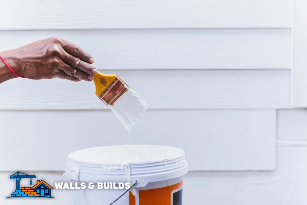 A hand holds a paintbrush dipped in white paint, brushing onto the rim of a paint bucket with orange paint visible, against a light-colored wall background.