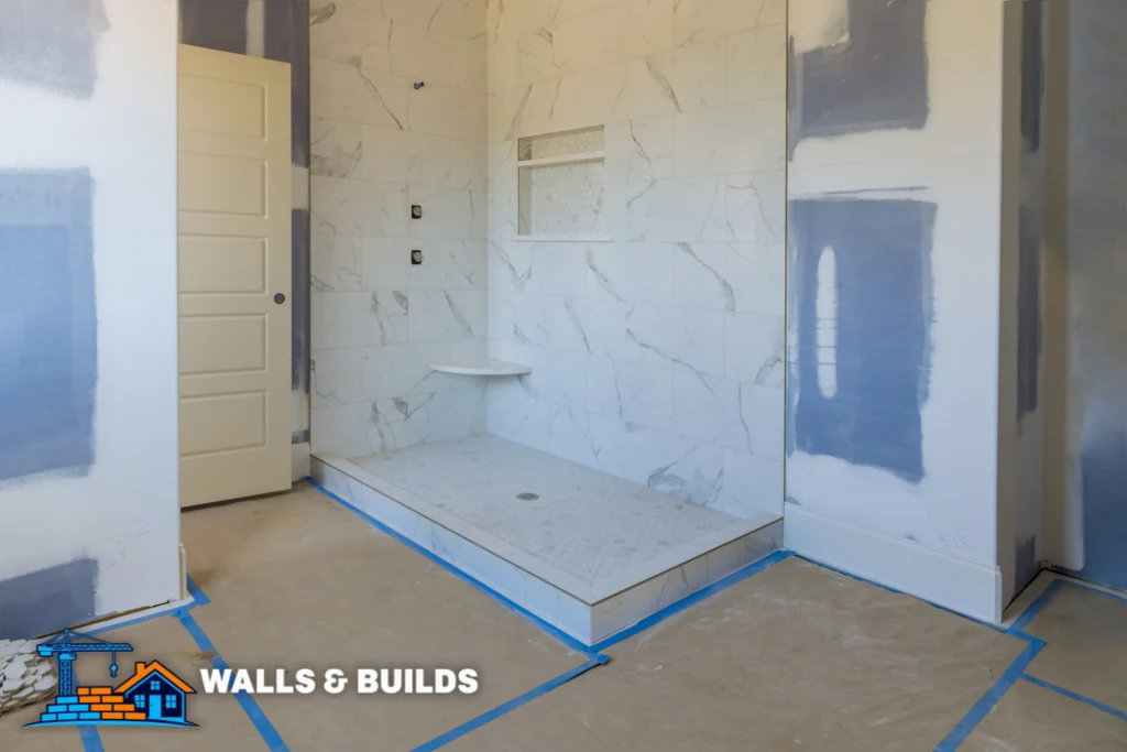 Interior shower area with a marble-tiled back wall, a built-in rectangular bench, and a glass panel; blue painter’s tape marks the floor layout and a white door stands on the left.