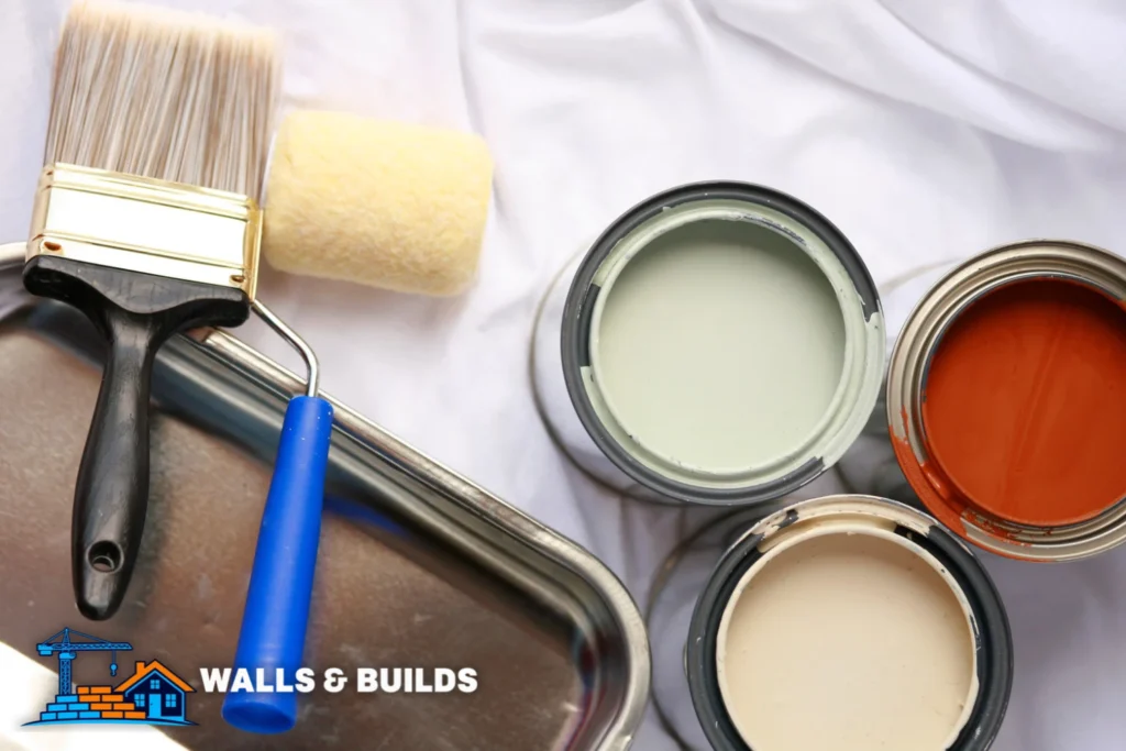 Paint tray with a blue-handled roller brush, and three open paint cans showing pale green, off-white, and orange paint on a white cloth.